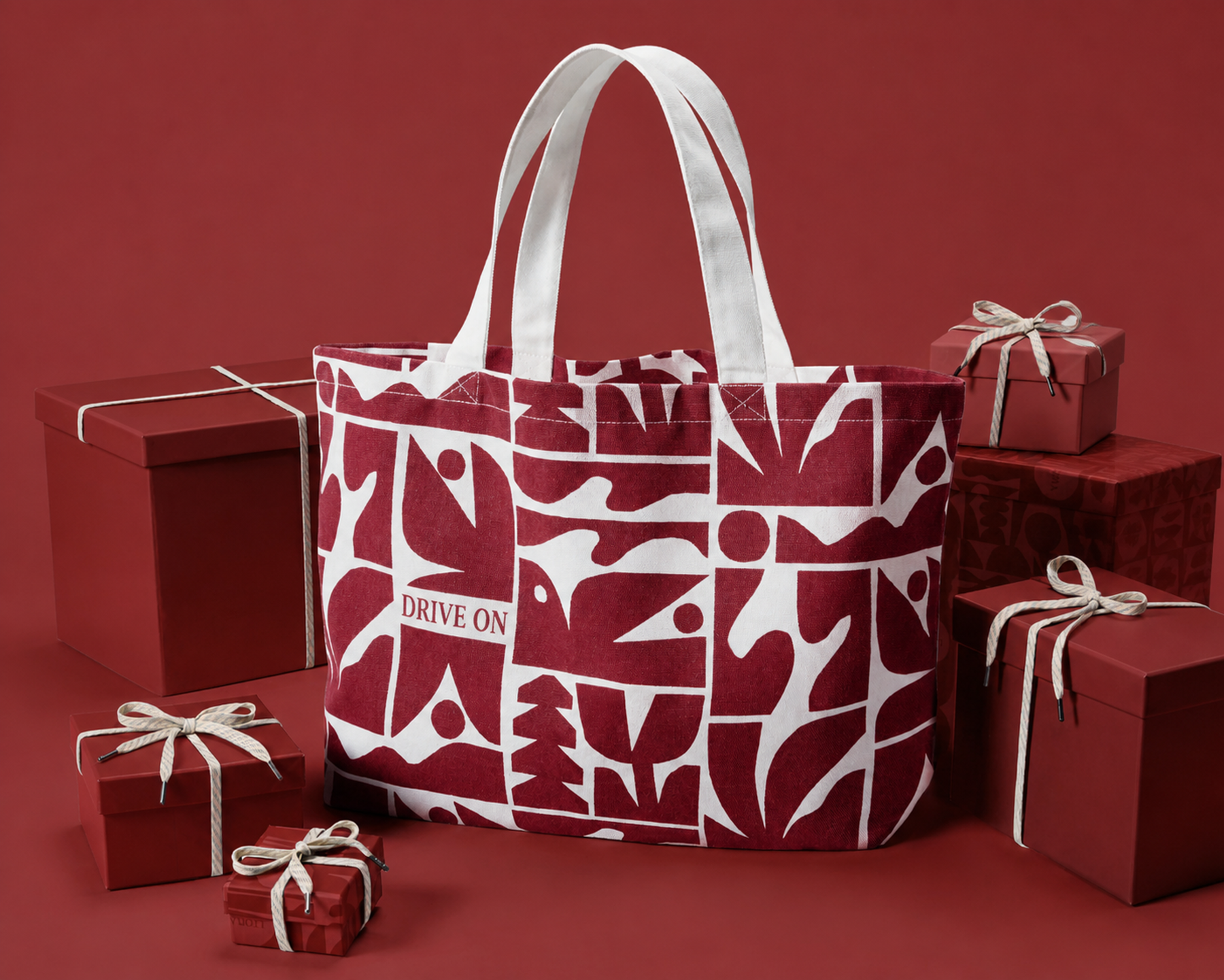 Red and white patterned DRIVE ON tote bag beside wrapped gift boxes on a red backdrop