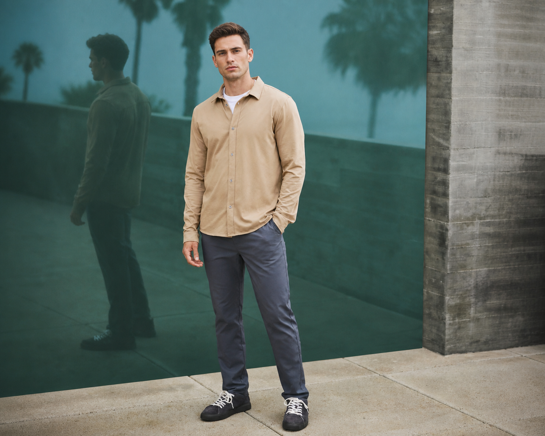 Man in a tan long-sleeve shirt and navy trousers standing against a teal wall