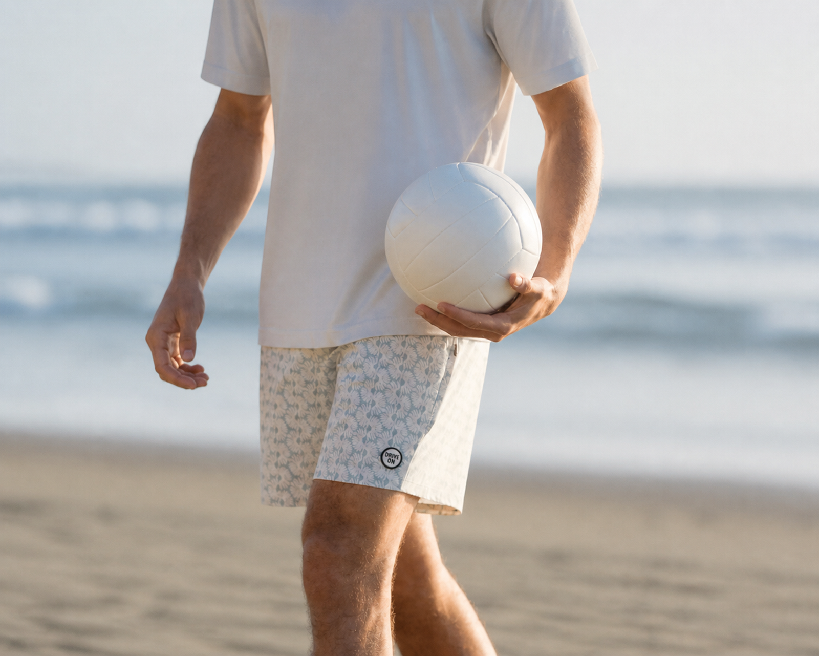 Man in a white tee and patterned shorts with a DRIVE ON hem patch, holding a volleyball on the beach