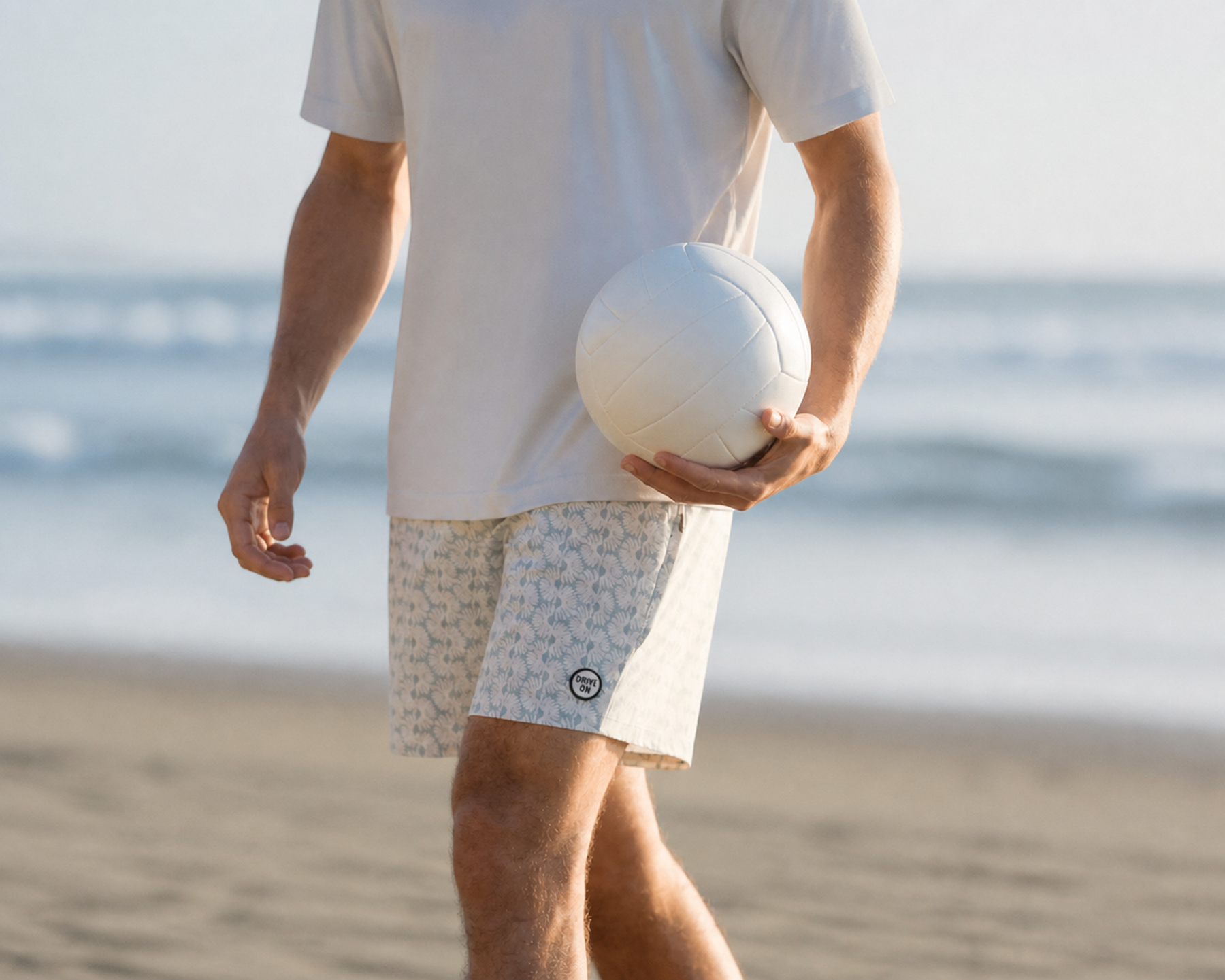 Man in a white tee and patterned shorts with a DRIVE ON hem patch, holding a volleyball on the beach
