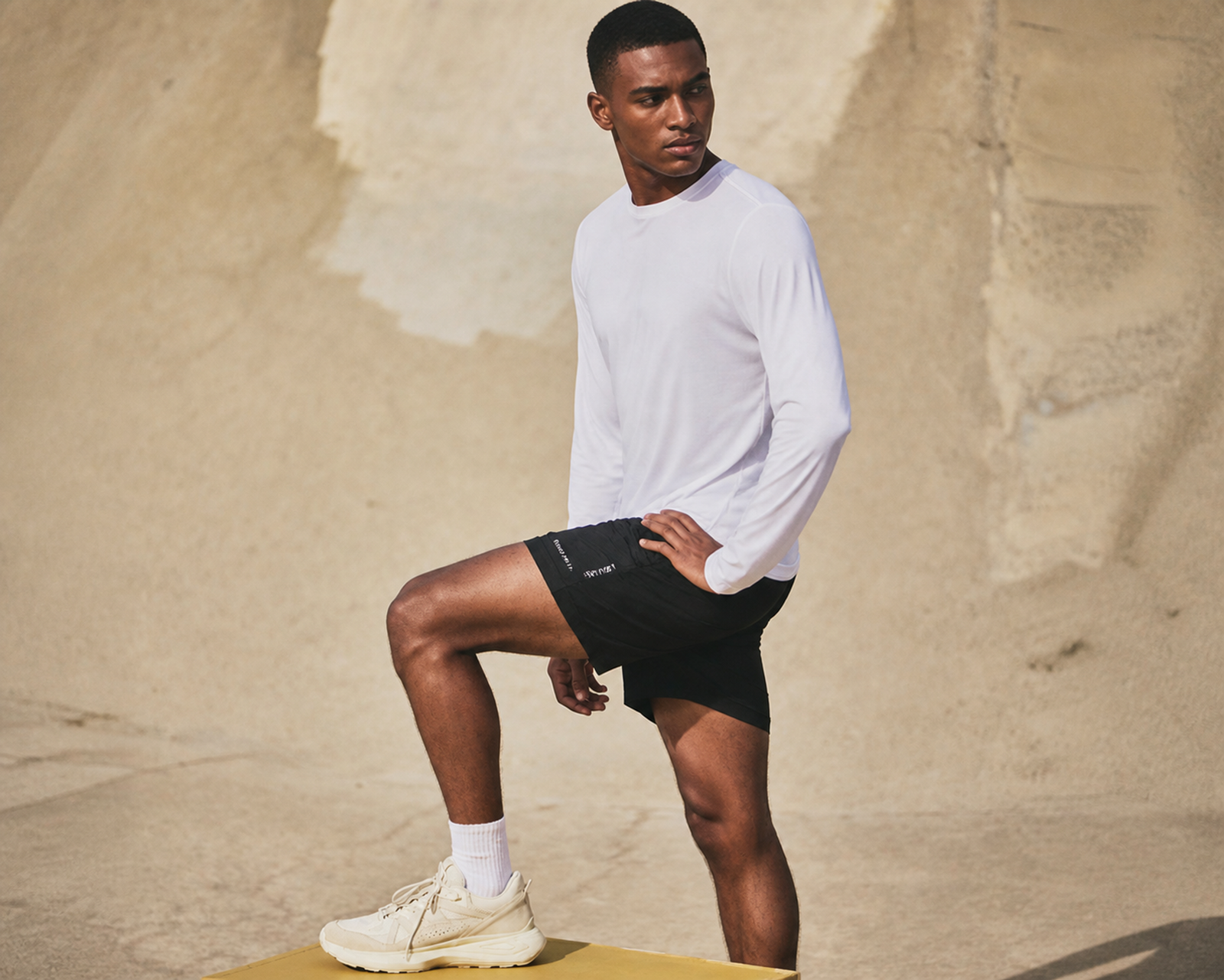 Man in a white long-sleeve top and black shorts with one foot propped on a block, warm stone backdrop
