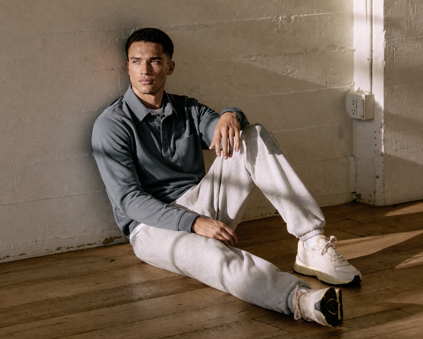 Man in a grey hoodie and joggers sitting cross-legged on a wood floor