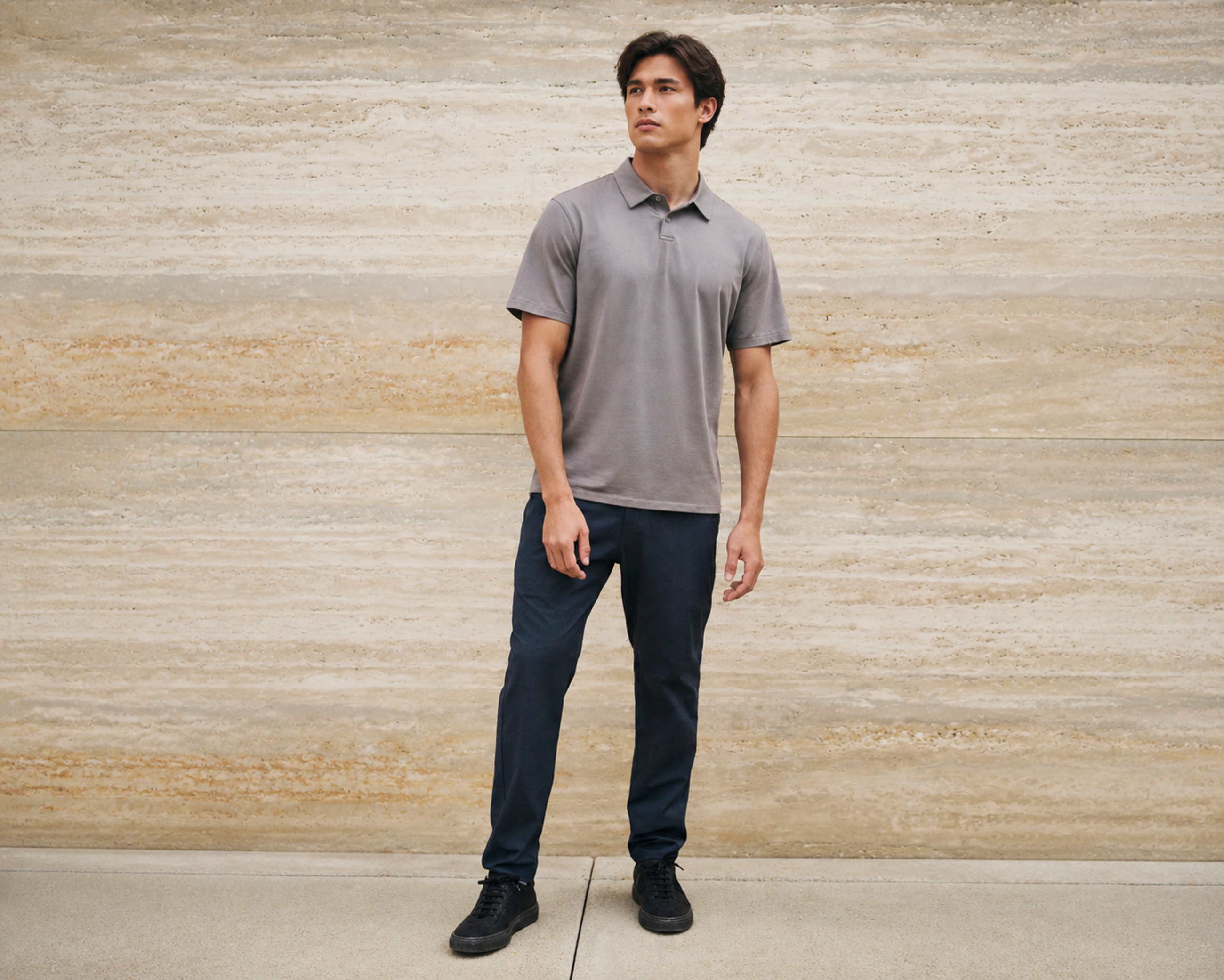 Man in a grey polo shirt and navy trousers standing against a warm travertine stone wall