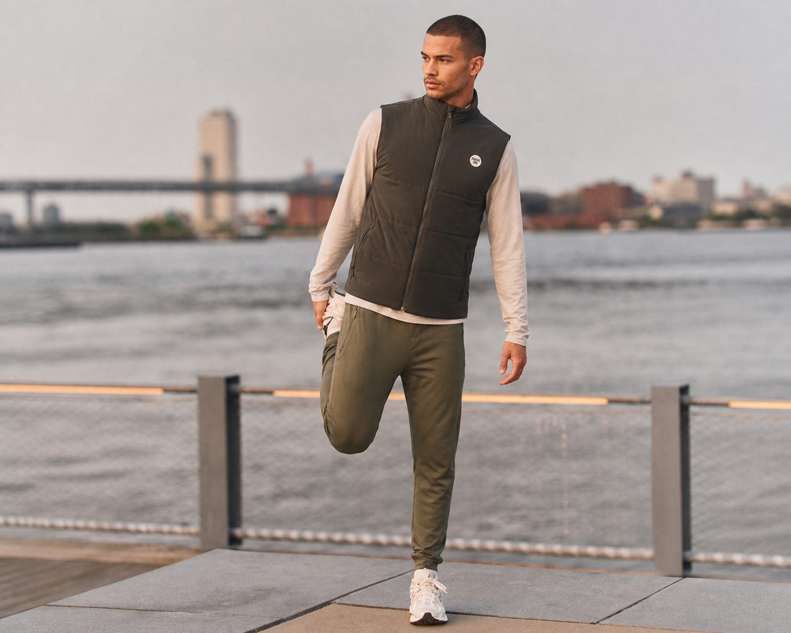 Man in an olive vest layered over a cream long-sleeve top and olive pants, stretching beside a waterfront