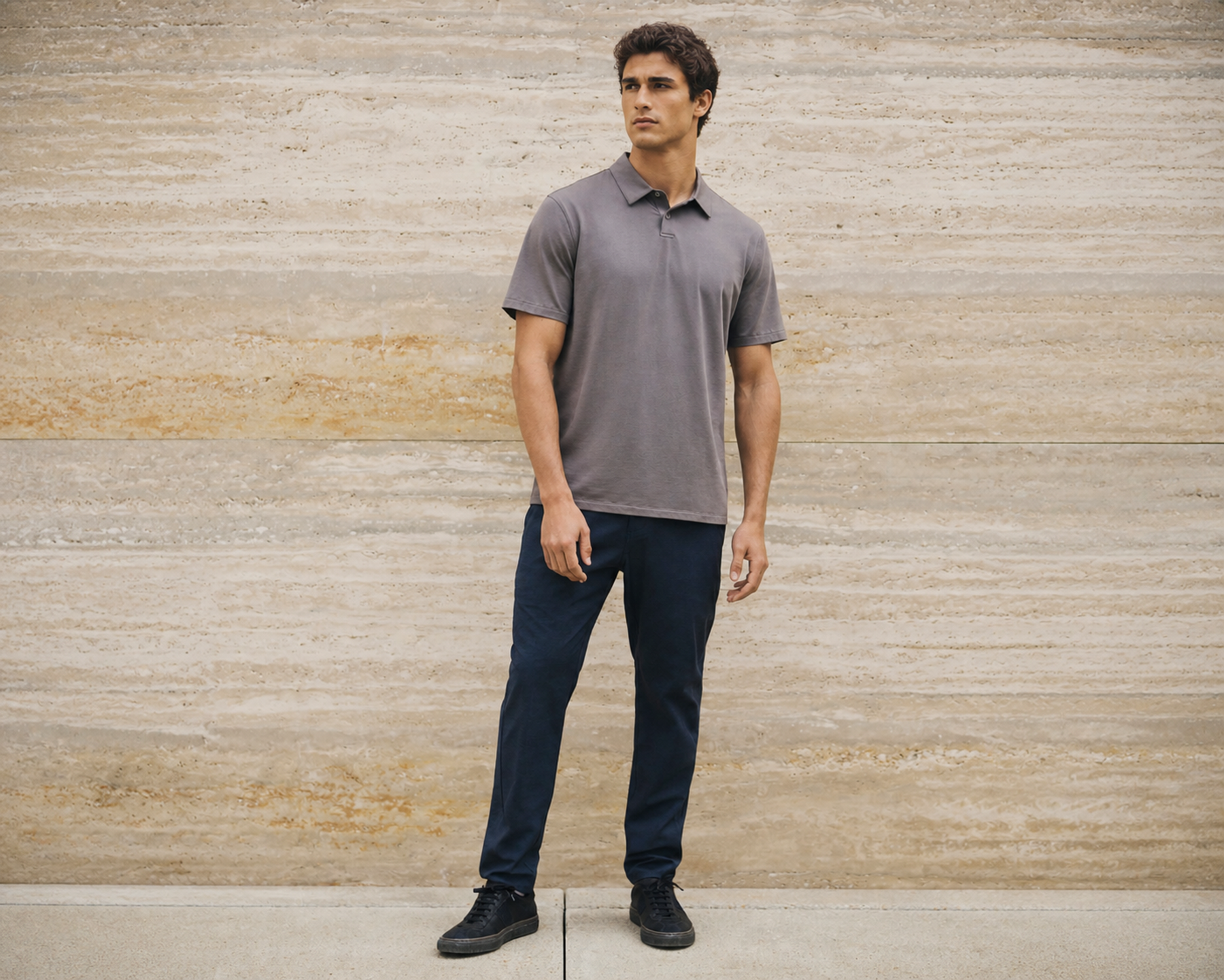 Man in a grey polo shirt and navy trousers standing against a travertine stone wall