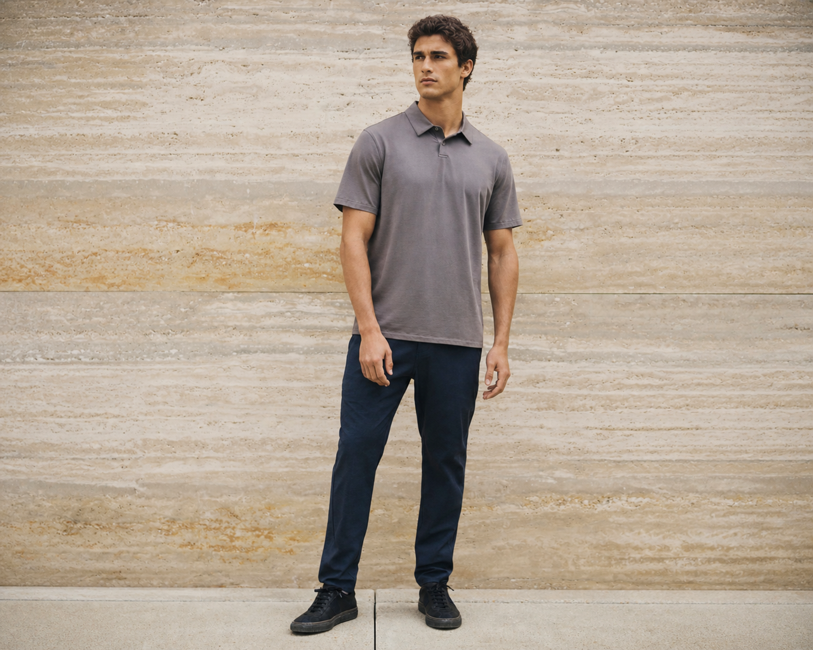 Man in a grey polo shirt and navy trousers standing against a travertine stone wall