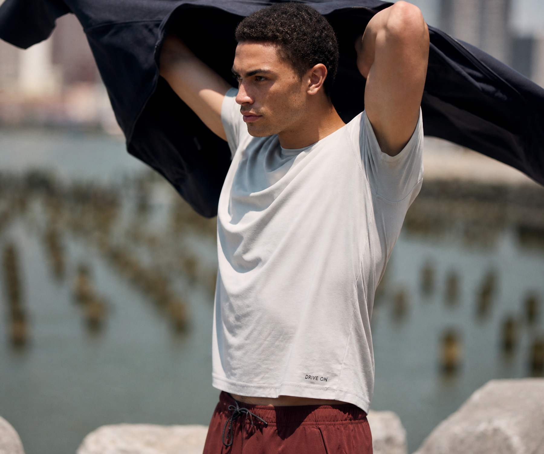 Man in a light grey DRIVE ON tee and maroon shorts, wind catching a dark jacket overhead