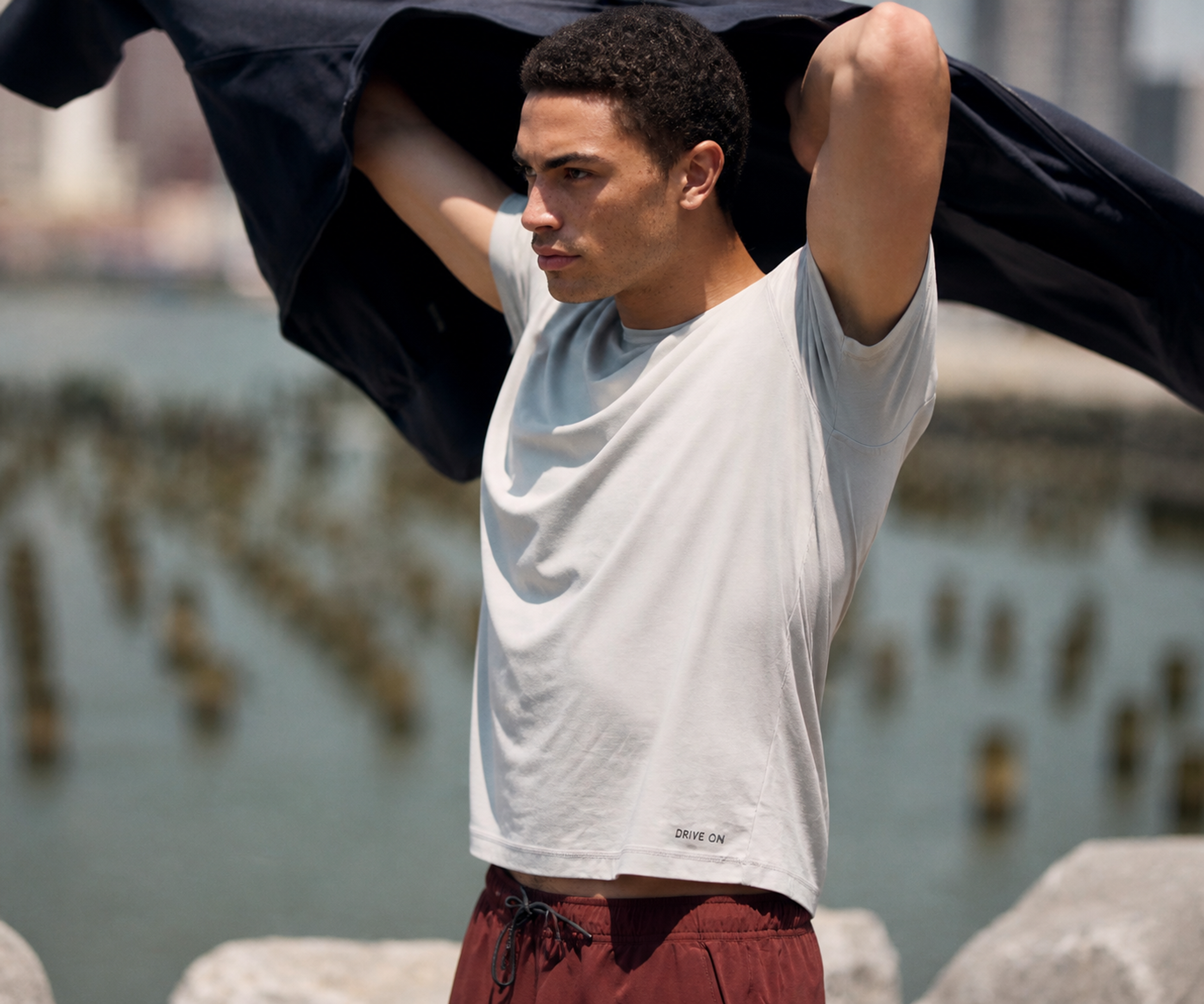 Man in a light grey DRIVE ON tee and maroon shorts, wind catching a dark jacket overhead