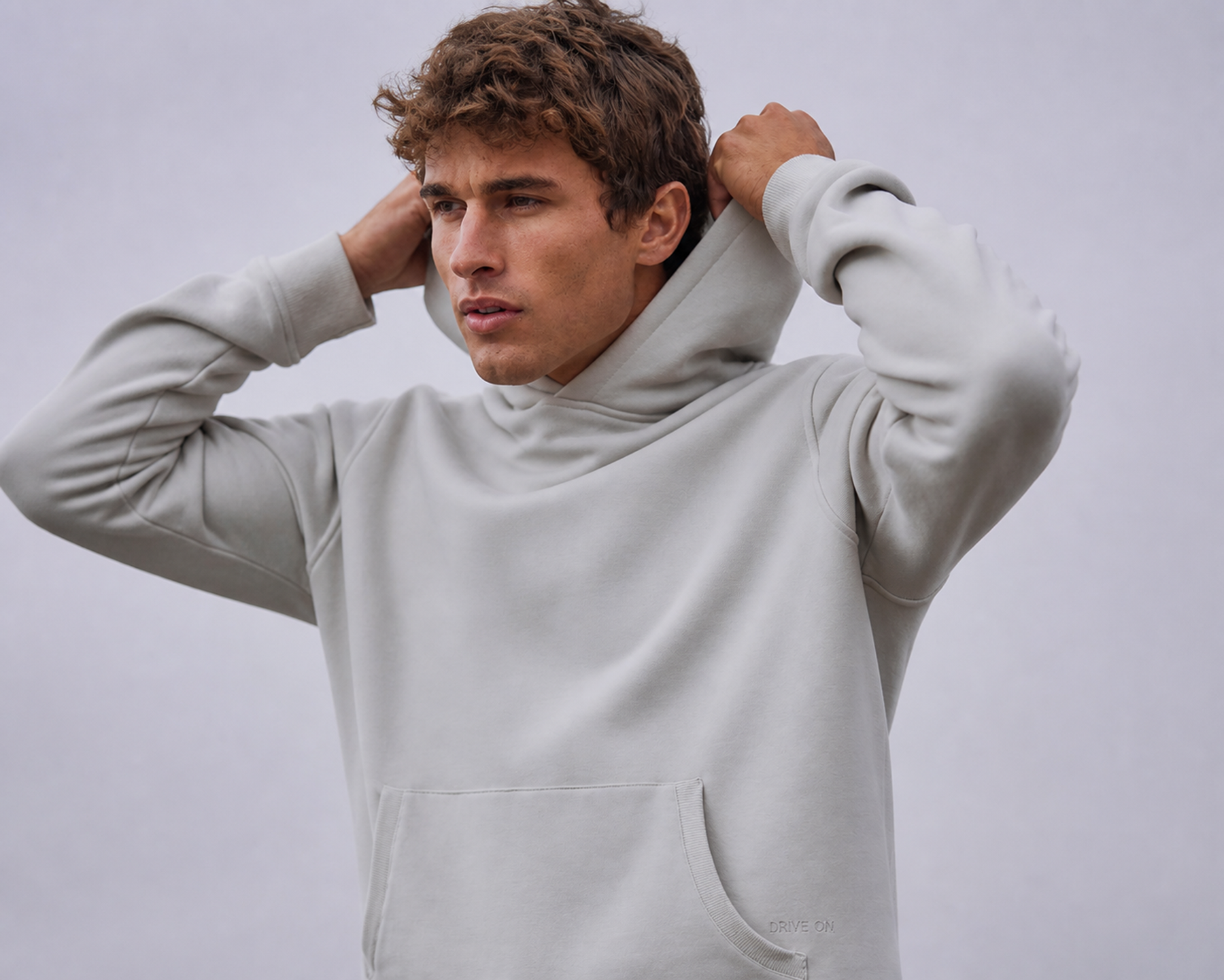 Man in a light grey hooded pullover adjusting his hood against a plain backdrop