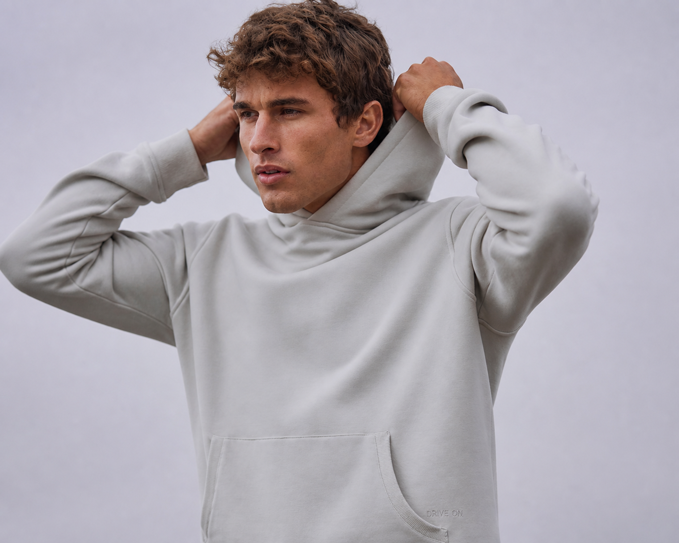 Man in a light grey hooded pullover adjusting his hood against a plain backdrop
