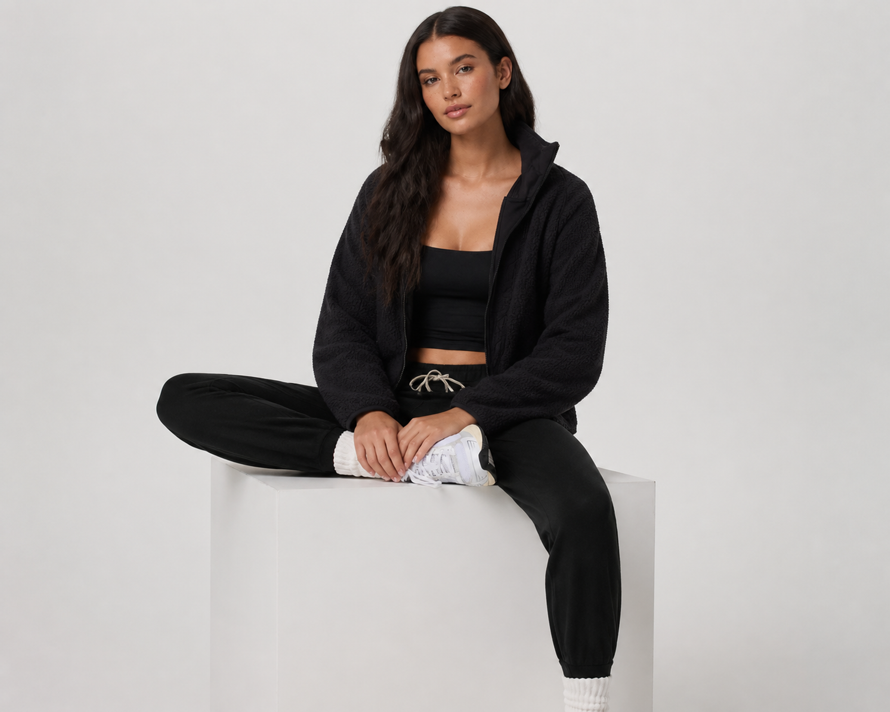 Woman in a black fleece jacket and joggers seated cross-legged on a white block