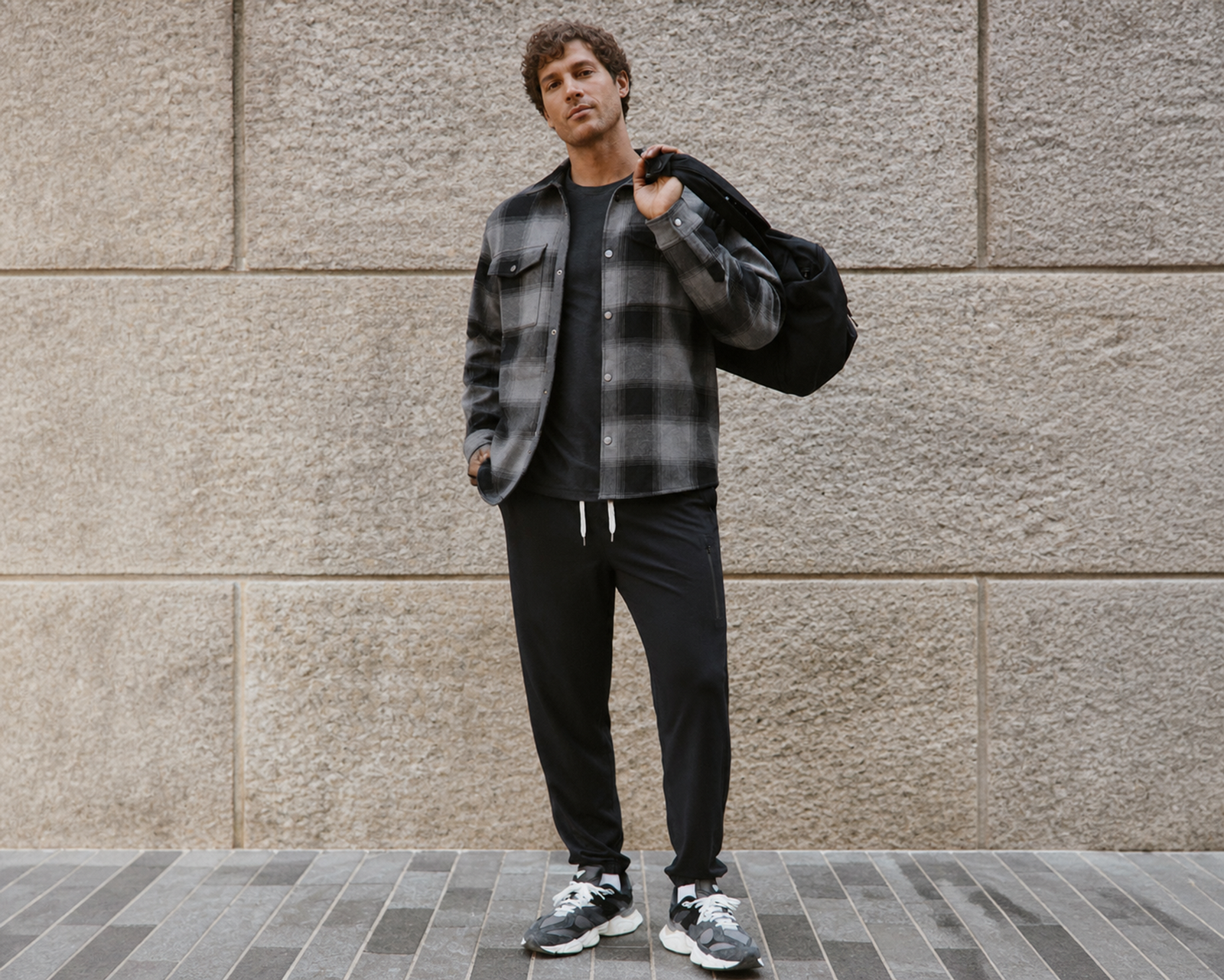 Man in a black and grey flannel-style shacket and joggers, carrying a bag over his shoulder on a city sidewalk