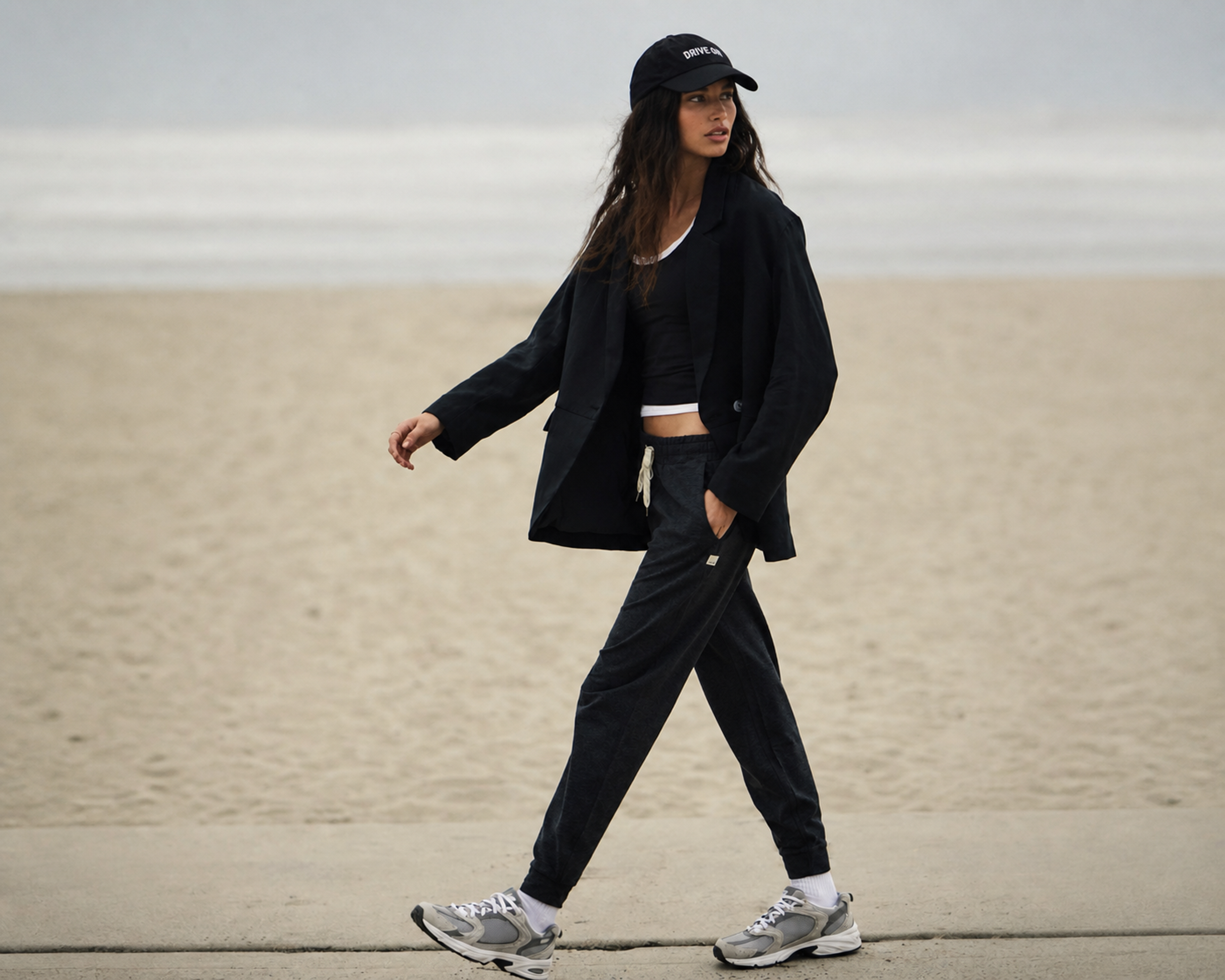 Woman in a black blazer, top and joggers walking along a sunlit beach boardwalk