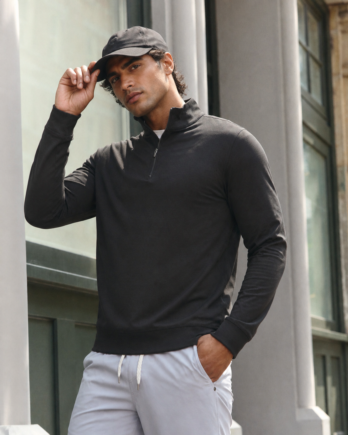 Man in a dark grey quarter-zip pullover and light grey shorts, tipping his cap, standing beside a glass storefront