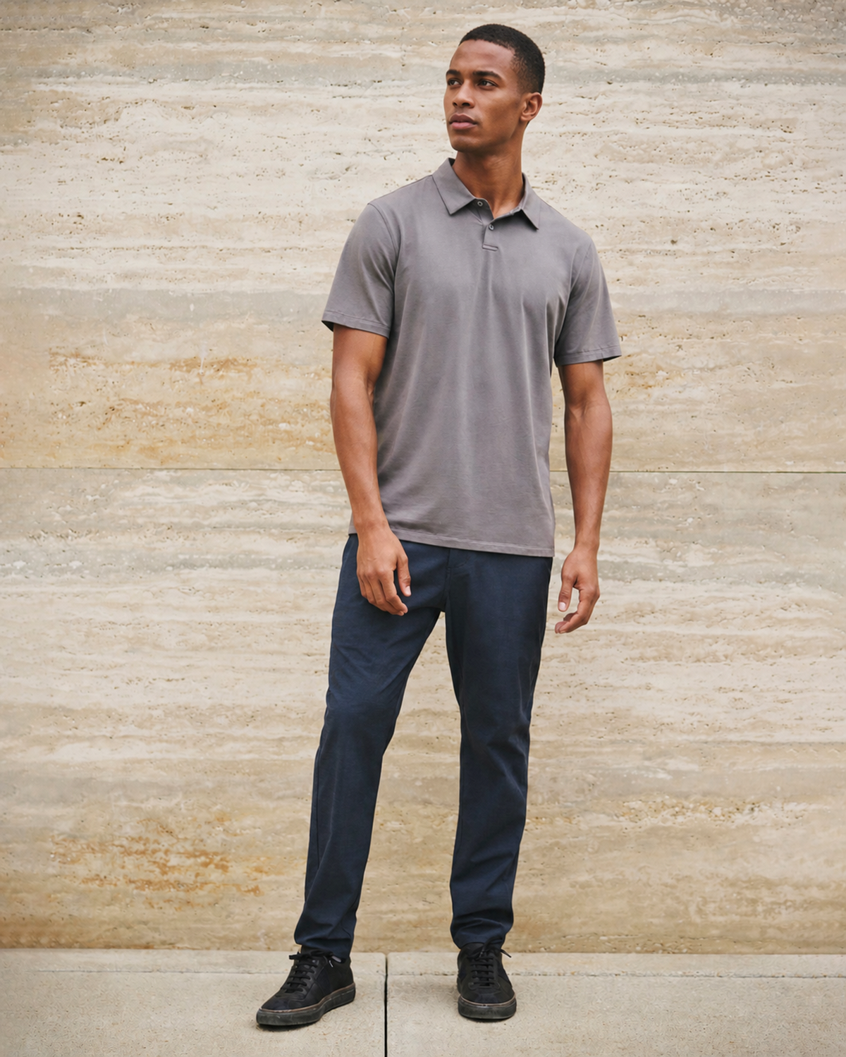 Man in a grey polo and navy trousers standing against a travertine stone wall