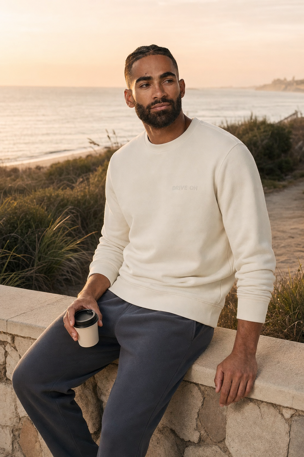 A man sitting on a wall with a coffee, beach behind, in the cream crewneck