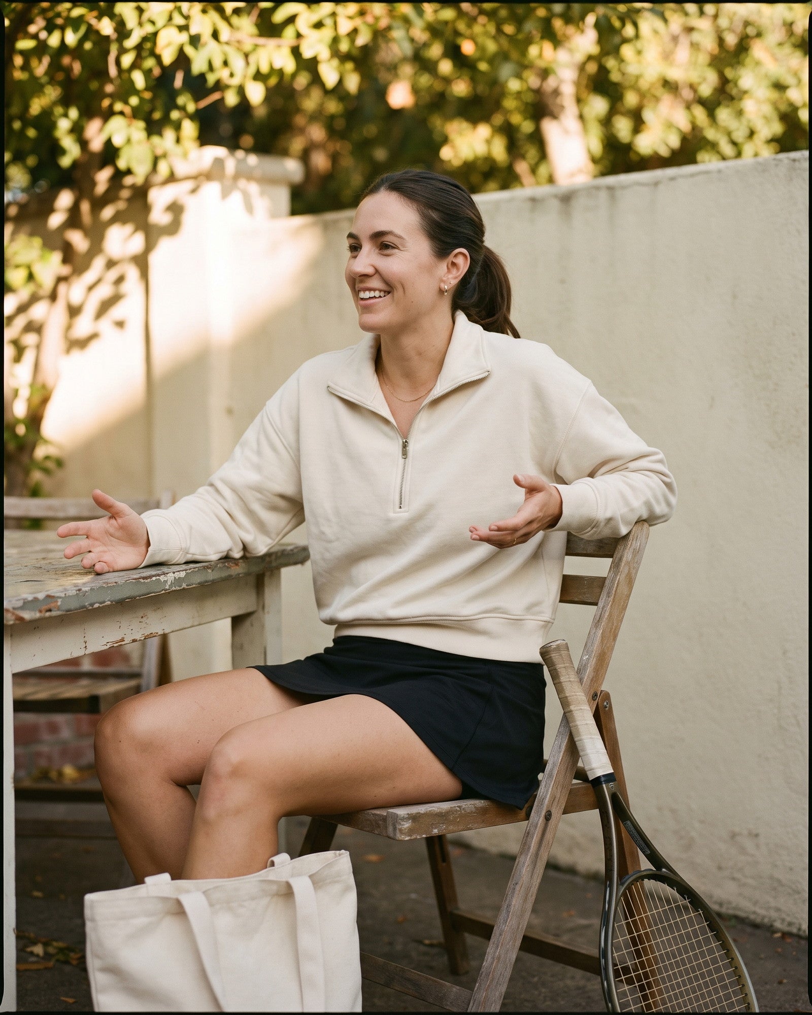 A woman at an outdoor table in the cream quarter-zip, tote and racquet beside the chair