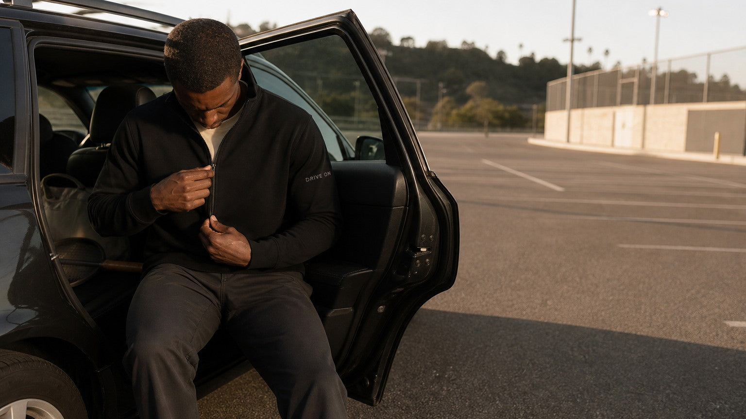 A man sitting in an open car door at the end of the day, drawing the black quarter-zip up