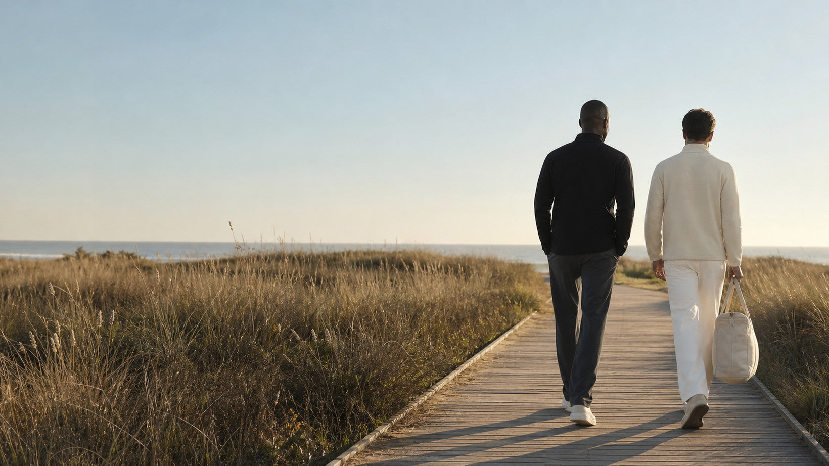 Two people walking away along a boardwalk through coastal dune grass into low sun